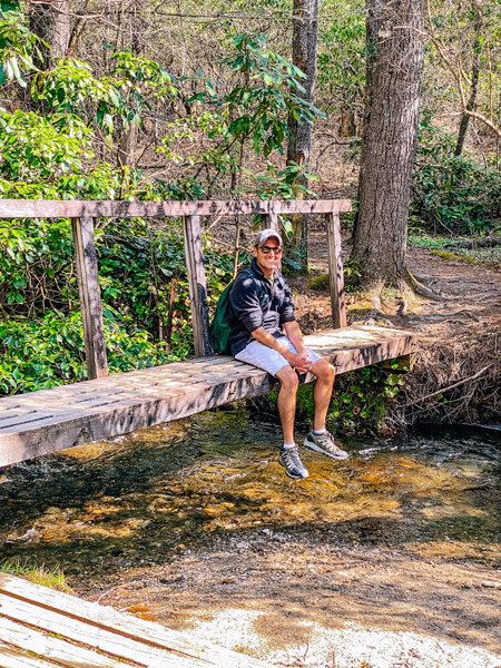 Pink Beds Loop Bridge with white brunette male sitting on the bridge over running stream. He is wearing shorts and a black fleece with a hat and sunglasses