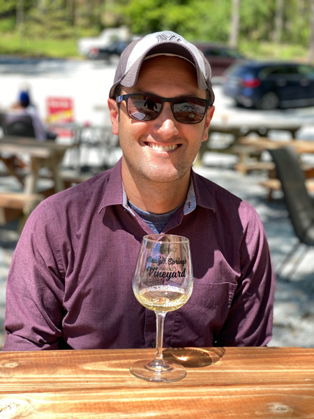 Sawyer Springs Vineyard Near Asheville portrait of white male wearing sunglasses and hat in purple shirt with white wine tasting glass