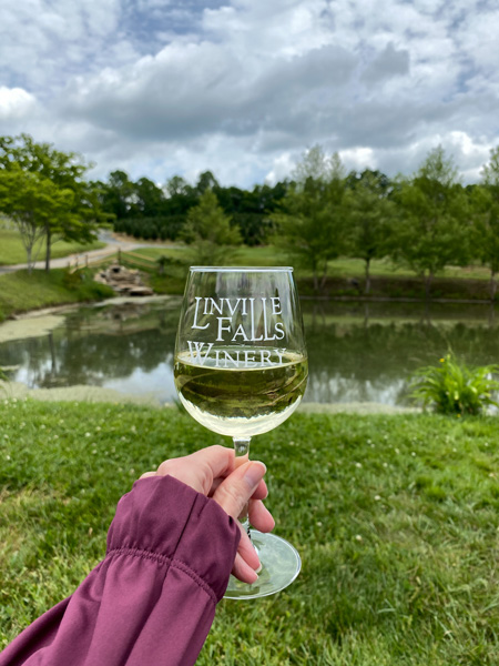 Linville Falls Winery with white hand in purple shirt raising white wine glass in front of pond