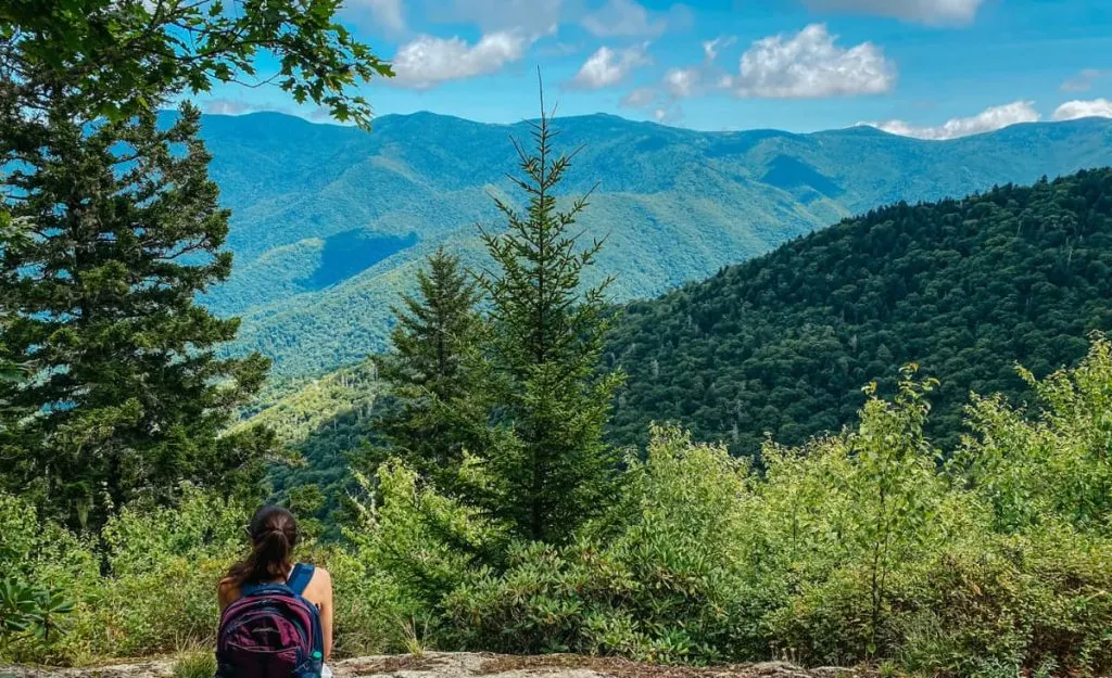 Little Butt Big Butt Trail NC with white brunette woman sitting on a rock with backpack looking out at the Black Mountains with trees and clouds in sky