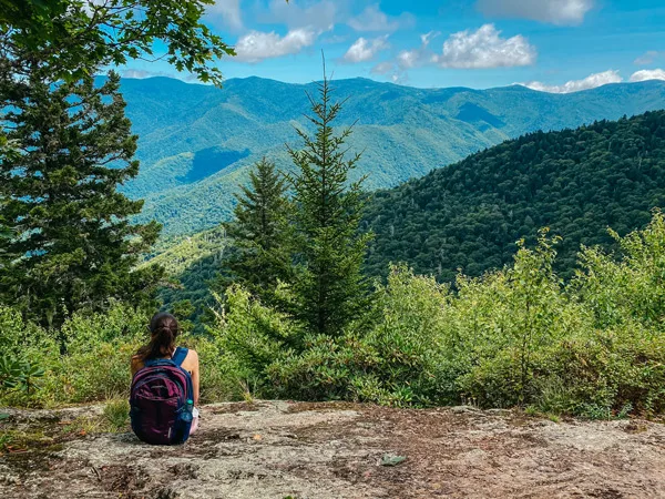 Little Butt Big Butt Trail Overlook with white brunette woman sitting on a rock with backpack looking out at the Black Mountains with trees and clouds in sky