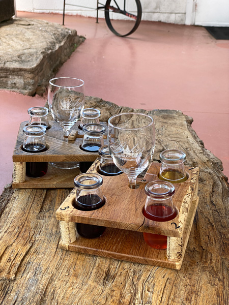 Wine Flight Baker Buffalo Creek Vineyard and Winery with four small glass milk bottles filled with wine and wine glass in middle of flight on wooden tree table