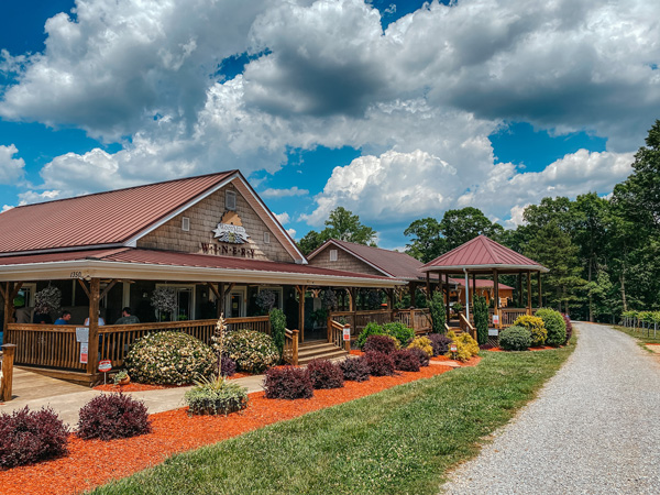 Woodmill Winery NC one story tasting room with gravel road, vineyards, and tables out front