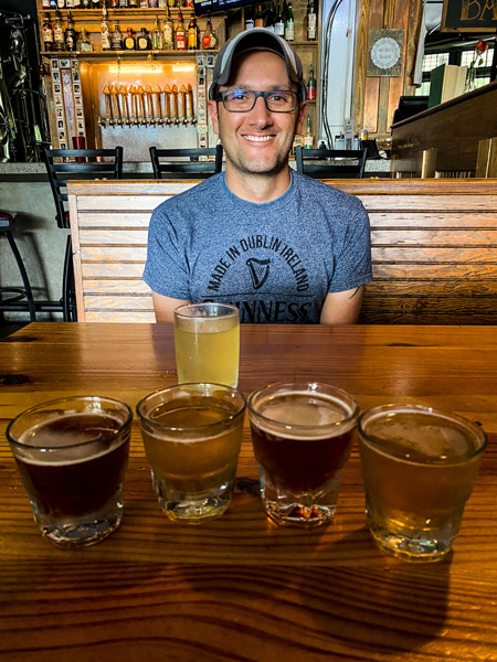 All Sevens Brewing Asheville NC with white brunette male in blue t-shirt with 5 flight glasses filled with various amber and blonde beers