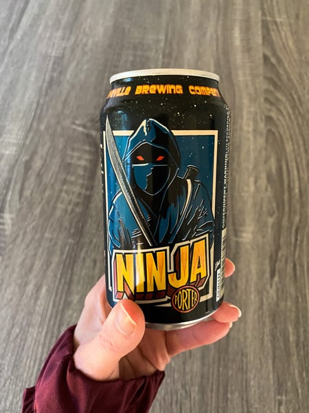 Asheville Brewing Canned Beer with white hand holding Nina Porter with illustration of person in armor with a sword in blue on can