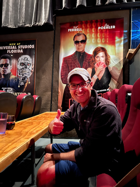 Asheville Pizza and Brewing in Asheville NC Theater with white brunette male in hat, fleece, and shorts sitting in movie theater seat with movie posters behind him and drinks on table in front of him