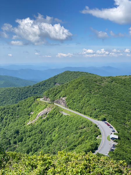 View of the Blue Ridge Parkway from Craggy Pinnacle (near Asheville, North Carolina) in the summer with green grass, view of Craggy Gardens Visitor Center parking lot, BRP road, and mountains around Craggy Gardens with blue sky and clouds