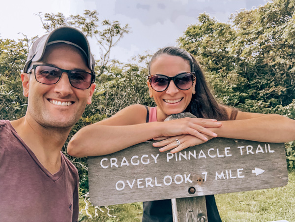 Craggy Pinnacle Trailhead with white brunette male and female with sunglasses in hiking clothes at trailhead sign that says Craggy Pinnacle Trail overlook .7 mile