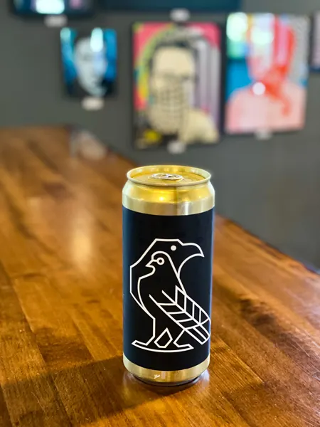 Eurisko Beer Company Asheville NC with can of beer with bird logo on label on brown table with pictures on wall blurred in background