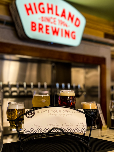 Highland Brewing Asheville NC with flight of blonde, stout, and amber beers, on a bar in front of Highland Brewing sign