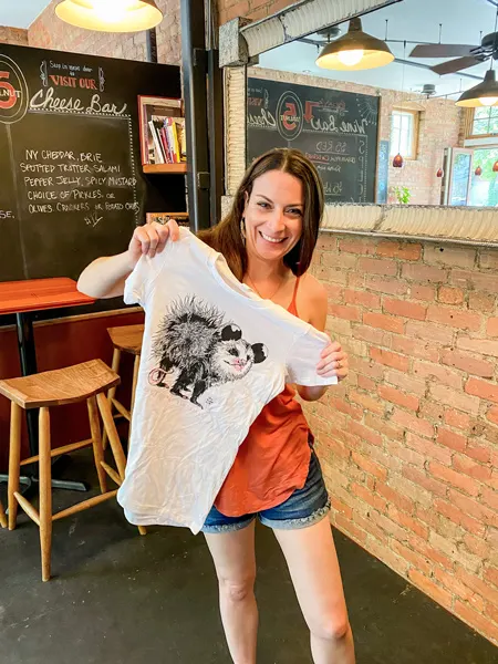 Izzys Coffee Asheville NC Shirt with white brunette female holding up shirt with opossum printed on it