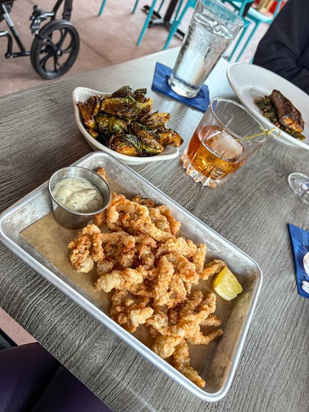 Fried Clams with tarter on metal tray and bowl of Brussels sprouts, glass of water, and lowball brown cocktail in the background at Jettie Rae's Oyster House in Asheville, NC 