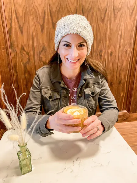 Latte Rowan Coffee Asheville NC with white brunette female in white hat, purple shirt, and green jacket drinking latte