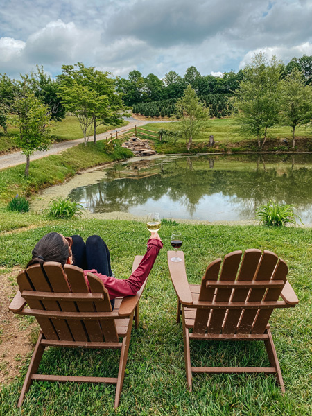 Linville Falls Winery with white brunette woman sitting in chair in front of vineyards and pond with glass of white wine