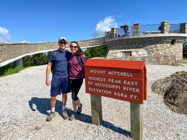Mount Mitchell Blue Ridge Parkway NC with white brunette male and female standing in front of sign that says highest peak east of the Mississippi River elevation 6684 feet with stone viewing platform behind them