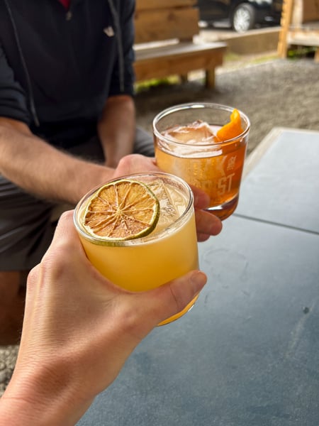 Oak and Grist Distillery Black Mountain NC with white male and female clinking glasses of orange and yellow cocktails with lime garnishes over a table