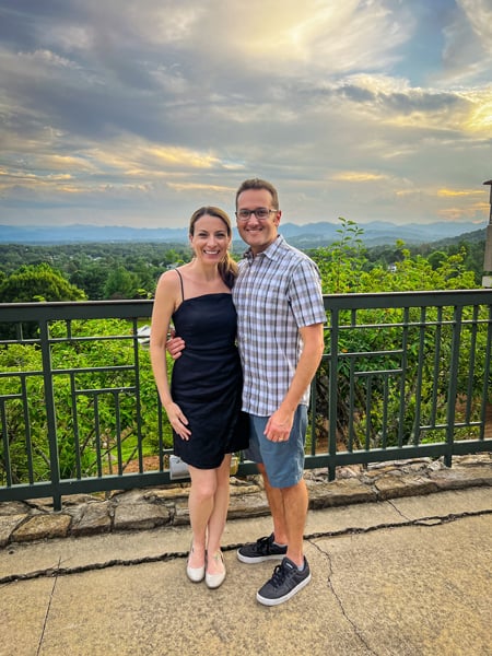 Christine and Tom, a white brunette female in black dress next to white brunette male in shorts and plaid shirt, with Blue Ridge Mountains in the background at The Omni Grove Park Inn in Asheville NC during the sunset