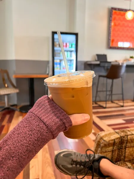 Penny Cup Coffee Shop in Asheville, North Carolina with white hand wearing purple sweater holding up a to-go iced coffee in front of table and walls