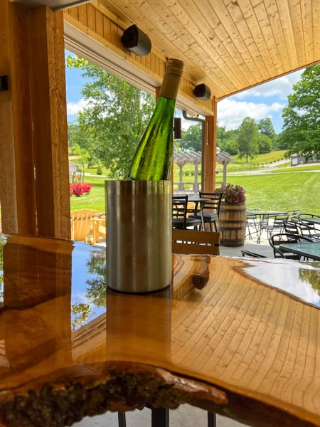 Souther Williams Vineyard Tasting Room with green bottle of white wine in chiller on wooden table in shaded tasting room overlooking green vineyard with pink flowering bushes