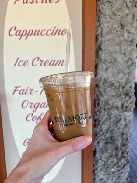 The Creamery at Biltmore Estate in Asheville NC Coffee in to-go plastic container held up by white hand in front of sign with menu items like ice cream and cappuccino