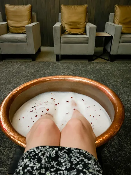 Wake Foot Sanctuary Asheville NC Spa Foot Soak with white legs in a big tub of water with rose petals and bubbles and gray armchairs with gold pillows in the background