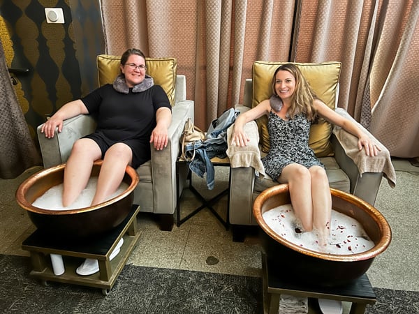 Christine and her friend, two white women in dresses, sitting in armchairs with foot soak tubs in front of them at Wake Foot Sanctuary Day Spa in Asheville, NC