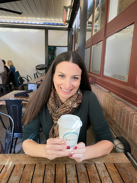 Christine, a white brunette woman sitting at table with to-go coffee cup from Well Bred Bakery at Corner Kitchen in Historic Biltmore Village in Asheville, NC