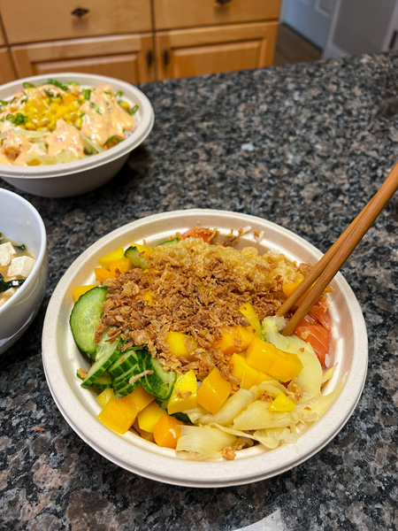 Yum Poke Spot Takeout in Asheville NC with takeaway bowl filled with white rice, mango, salmon, fried onions, and cucumbers with chopsticks