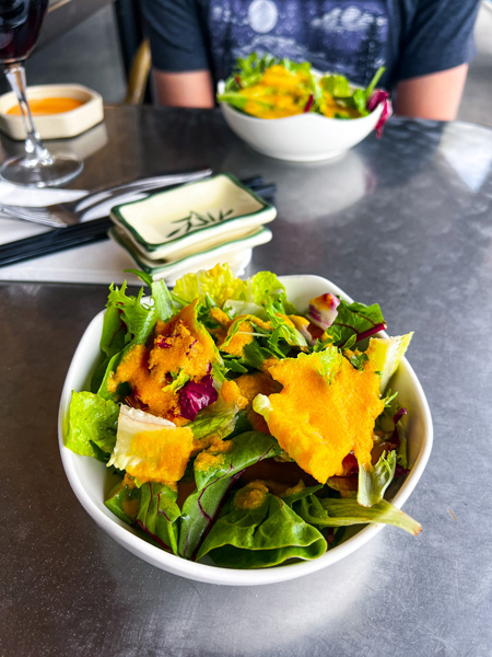 Zen Sushi in Asheville, NC Ginger Salad with bright green lettuce and orange colored ginger dressing on silver table with second salad and empty stacked soy sauce bowls in the back