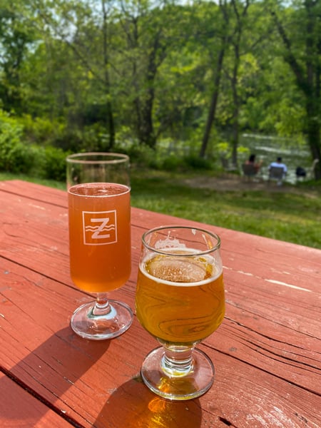 Two beers, one yellow and one light amber/orange, on picnic table with green grass and people in chairs in front of French Broad River in the background at Zillicoah Beer Company