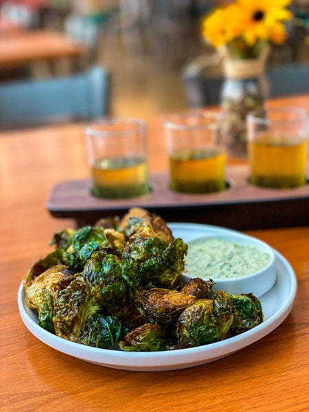 Brussel Sprouts Noble Cider Asheville on white plate with sauce, cider flight in background, and yellow flowers on table