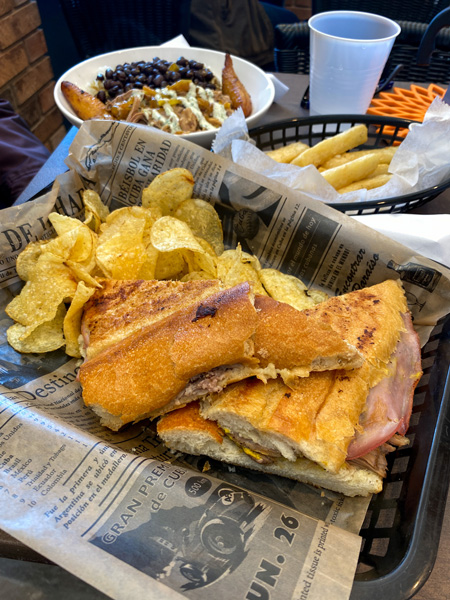 Cousins Cuban Cafe Cuban sandwich with pork, pickles, and chips on cafe table with yucca fries and plantain bowl in background