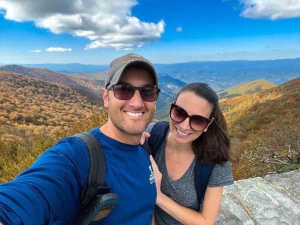 Craggy Gardens Visitor Center during the Fall in Asheville, NC with selfie of white brunette male with hat, blue shirt, and sunglasses, and white brunette female with sunglasses in gray t-shirt standing in front of overlook with yellow and red fall foliage and Blue Ridge Mountains in background