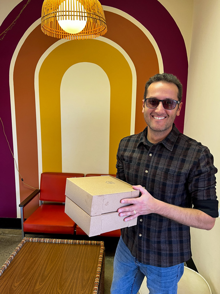 Dough House in Black Mountain, NC with white brunette male in jeans and sunglasses holding two boxes of takeaway donuts in front of red, orange, and yellow mural