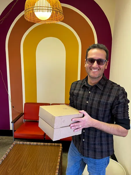 Dough House in Black Mountain, NC with white brunette male in jeans and sunglasses holding two boxes of takeaway donuts in front of red, orange, and yellow mural