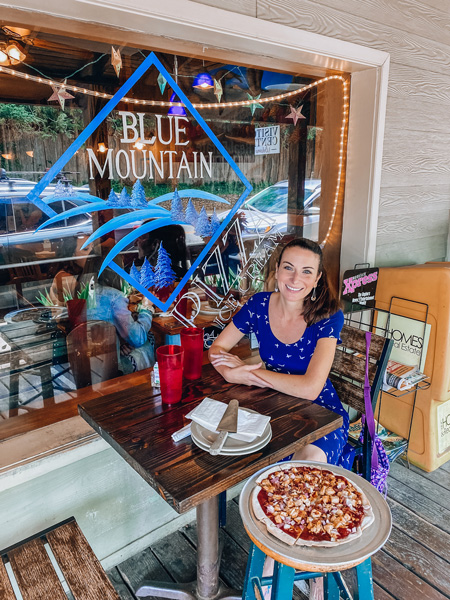 Gluten-free Blue Mountain Pizza in Weaverville NC with white brunette woman in blue bird dress sitting next to a gluten-free pizza and eating outside with Blue Mountain Pizza logo in background