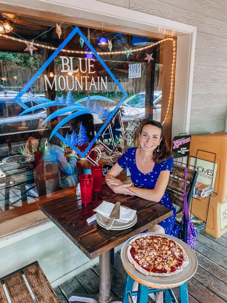 Gluten free Blue Mountain Pizza Weaverville NC with white brunette woman in blue bird dress sitting next to a gluten-free pizza and eating outside with Blue Mountain Pizza logo in background