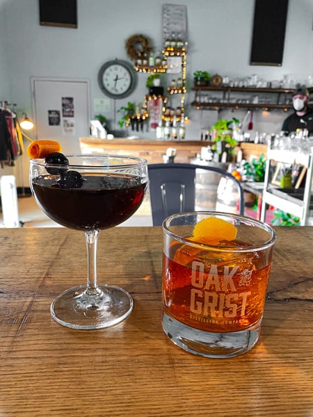 Oak and Grist Distilling Company low ball orange cocktail and martini like glass red cocktail on wooden table with bar blurred in background