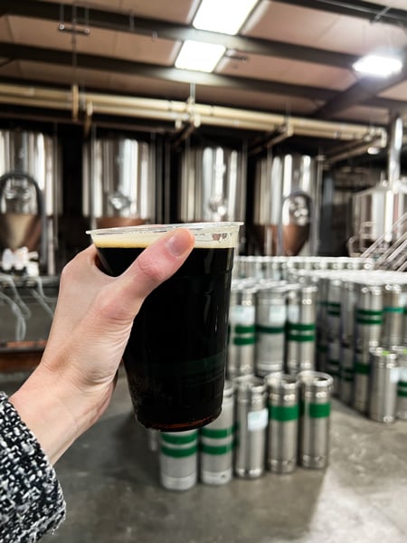 Pisgah Brewing Black Mountain Brewery with white female hand holding dark vanilla stout in a plastic cup in front of brewing steel containers
