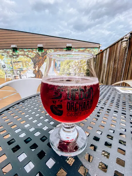 Urban Orchard Cider Asheville NC with red cider in glass on outdoor table with mural and cloudy sky in the background