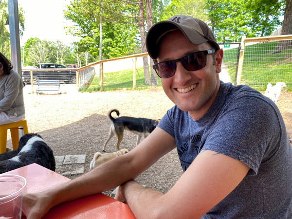 WagBar in Weaverville, NC with white brunette male wearing hat and sunglasses drinking a cider from plastic cup surrounded by dogs