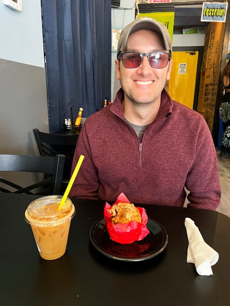 White brunette male with hat, sunglasses and maroon sweater with an iced coffee, and vegan, gluten-free muffin at Yellow Coffee Mug Lounge in Weaverville NC