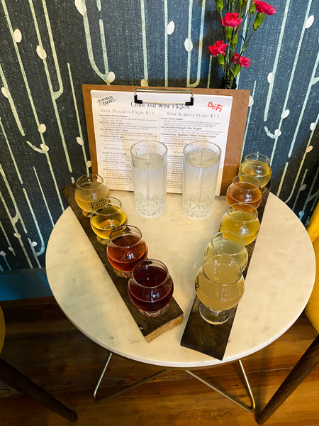 Botantist and Barrel Cider Asheville NC with two flights of cider of varying colors from yellow, gold, red, and orange on small table with menu in background