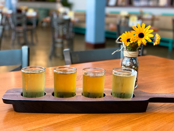 Flight Noble Cider Asheville NC with four light to yellow colored ciders in wooden flight with yellow flowers in vase on table