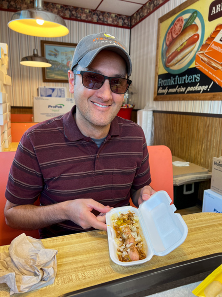 Hot Dog World Hendersonville NC with white male in maroon shirt with sunglasses and hat holding a bun-less hot dog with chili