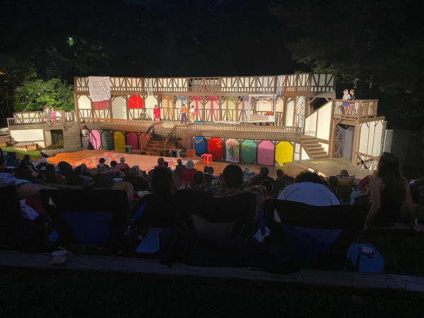 Montford Park Players Shakespeare in the Park with colorful open air theater lit up at night with rows of people in lawn chairs in front of it