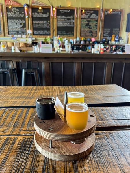Oklawaha Brewing Company Hendersonville NC with beer flight with two light beers and a dark beer on wooden table with taproom beer list blurred in background