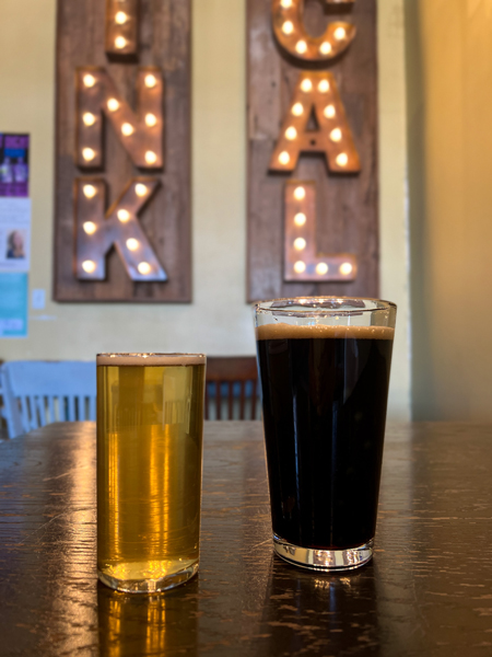 Oklawaha Brewing Hendersonville NC with light small beer and large dark beer on wooden table with lit up drink local sign on in the background