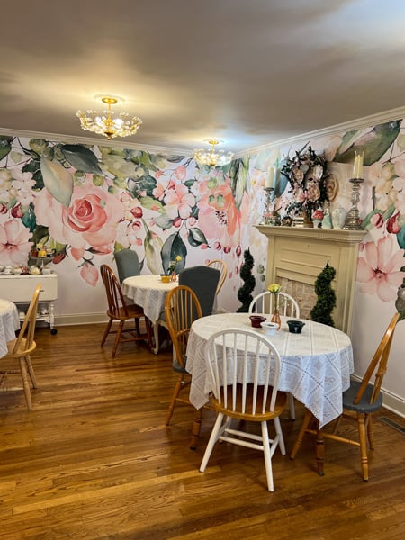 The Book and Bee Cafe and Tearoom dining room with white fireplace, flowered wallpaper, and tables with white tablecloths