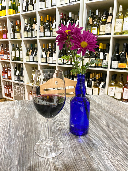 Wine Sage and Gourmet Hendersonville NC with glass of red wine on gray table with blue vase filled with pink flowers surrounded by shelves of bottled wine